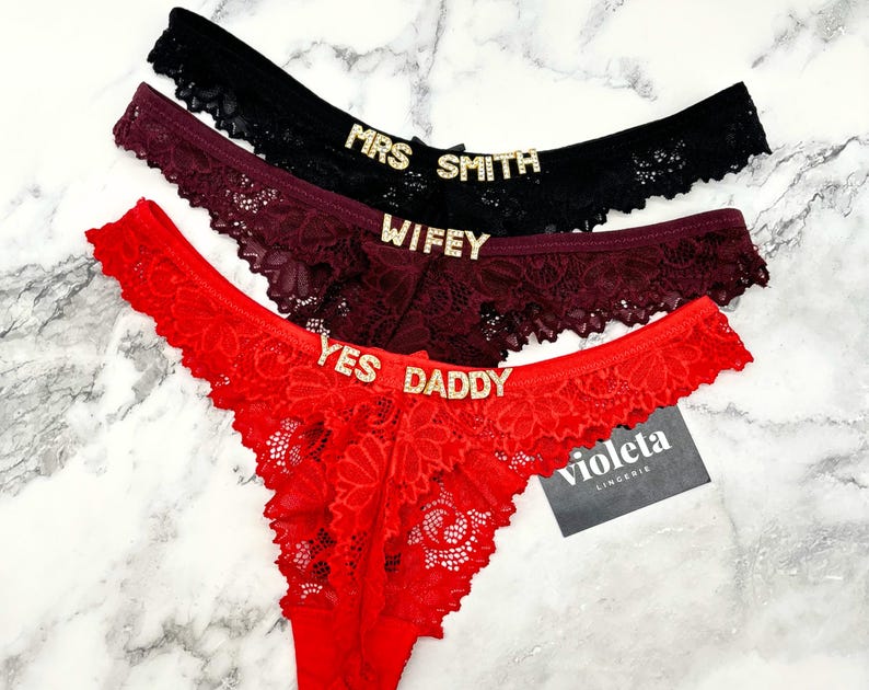 May include: Three lace thongs in black, burgundy, and red, each featuring gold-toned rhinestone lettering on the waistband. The black pair reads MRS SMITH, the burgundy pair reads WIFEY, and the red pair reads YES DADDY. A small black tag reads violeta LINGERIE.