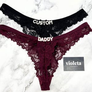 May include: Two pairs of lace thong underwear, one black and one burgundy, each featuring a silver-toned rhinestone embellishment on the front. The black pair displays the word CUSTOM, while the burgundy pair displays the word DADDY. A small black card with the white text violeta LINGERIE is included.