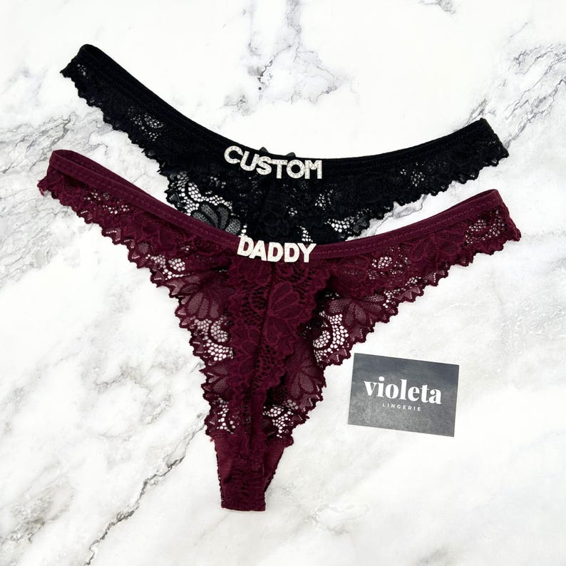 May include: Two pairs of lace thong underwear, one black and one deep burgundy. The black pair features the word CUSTOM in silver-toned rhinestone letters, while the burgundy pair features the word DADDY in silver-toned rhinestone letters.