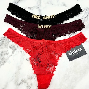 May include: Three lace thongs in black, burgundy, and red. The black pair features the text MRS SMITH in gold-toned letters, the burgundy pair features the text WIFEY in gold-toned letters, and a black tag with the white text violeta LINGERIE.
