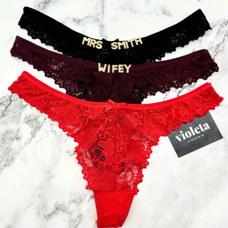May include: Three lace thongs in black, burgundy, and red. The black pair features the text MRS SMITH in gold-toned letters, the burgundy pair features the text WIFEY in gold-toned letters, and a black tag with the white text violeta LINGERIE.