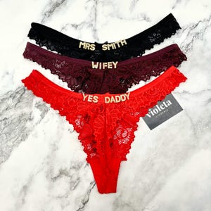 May include: Three pairs of lace thong underwear in black, burgundy, and red. Each pair features gold-toned rhinestone lettering on the front waistband. The black pair reads MRS SMITH, the burgundy pair reads WIFEY, and the red pair reads YES DADDY.