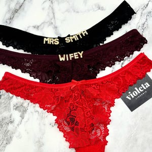 Può includere: Tre paia di biancheria intima in pizzo con colori e decorazioni diversi. Il paio superiore è nero con lettere dorate scintillanti che dicono "MRS SMITH". Il paio intermedio è bordeaux con lettere dorate scintillanti che dicono "WIFEY". Il paio inferiore è rosso con un motivo di pizzo floreale. Il paio rosso ha un'etichetta bianca che dice "violeta lingerie".
