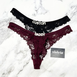 May include: Two lace thongs, one black and one burgundy, featuring silver-toned rhinestone lettering on the front. The black thong displays the word CUSTOM, while the burgundy thong displays the word DADDY. A small black card with the text violeta LINGERIE is included.