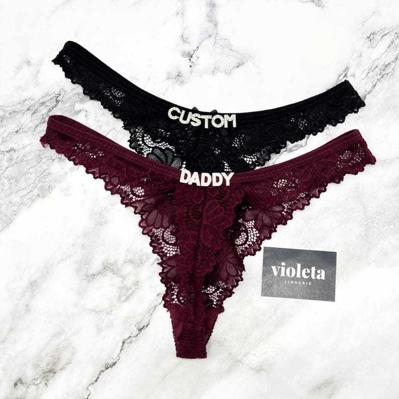 May include: Two lace thongs, one black and one burgundy, featuring silver-toned rhinestone lettering on the front. The black thong displays the word CUSTOM, while the burgundy thong displays the word DADDY. A small black card with the text violeta LINGERIE is included.
