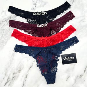 May include: Four lace thongs in black, burgundy, red, and navy blue. The black pair features the word CUSTOM in rhinestones, and the burgundy pair features the word DADDY in rhinestones. A small black card with the white text violeta LINGERIE is included.
