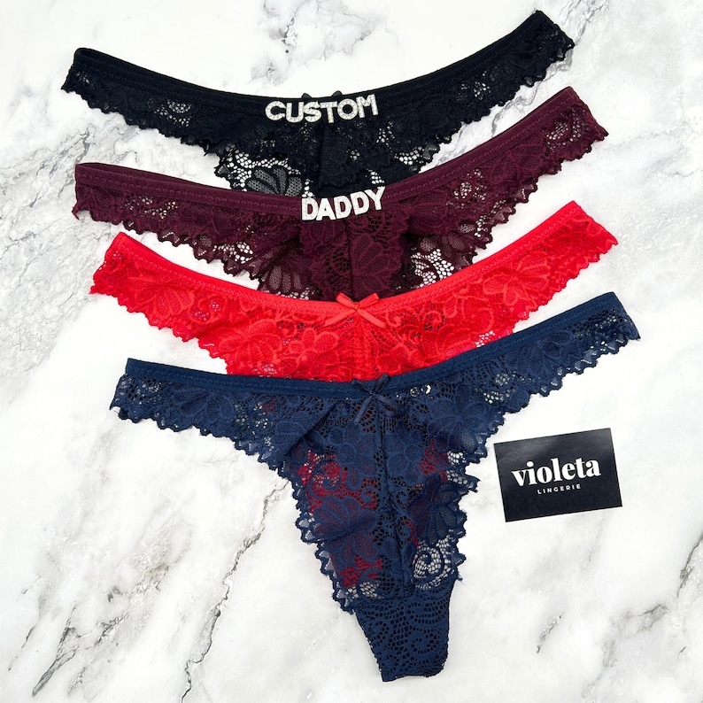 May include: Four lace thongs in black, burgundy, red, and navy blue. The black pair features the word CUSTOM in rhinestones, and the burgundy pair features the word DADDY in rhinestones. A small black card with the white text violeta LINGERIE is included.