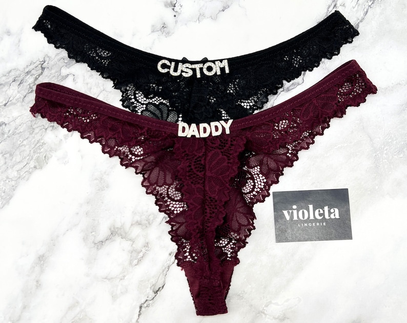May include: Two lace thongs, one black and one burgundy, featuring silver-toned rhinestone lettering. The black thong displays the word CUSTOM, while the burgundy thong displays the word DADDY. A small black card with the text violeta LINGERIE is included.