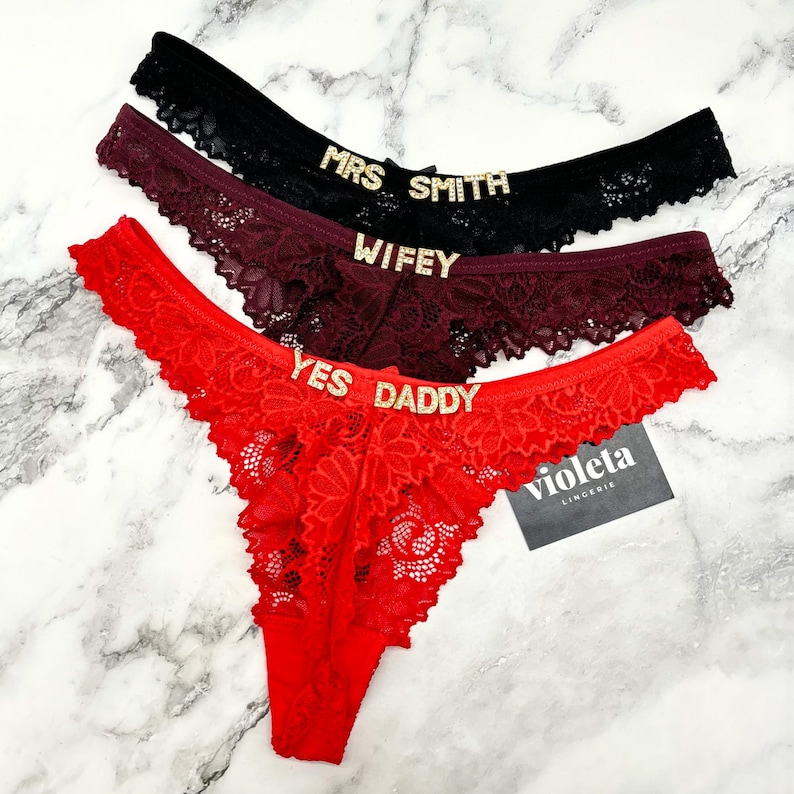 May include: Three pairs of lace thong underwear in black, burgundy, and red. Each pair features gold-toned rhinestone lettering on the waistband. The black pair reads MRS SMITH, the burgundy pair reads WIFEY, and the red pair reads YES DADDY.