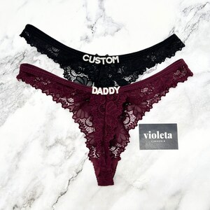 May include: Two lace thongs, one black and one burgundy, featuring rhinestone lettering on the front. The black thong displays the word CUSTOM, while the burgundy thong features the word DADDY. A small black card with the text violeta LINGERIE is included.