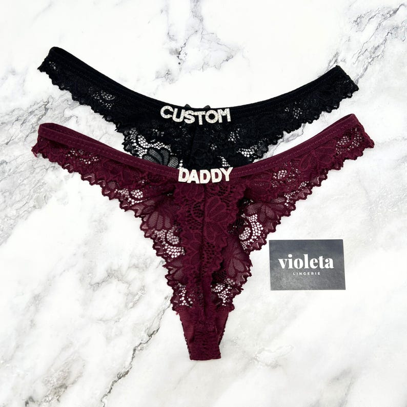 May include: Two lace thongs, one black and one burgundy, featuring rhinestone lettering on the front. The black thong displays the word CUSTOM, while the burgundy thong features the word DADDY. A small black card with the text violeta LINGERIE is included.