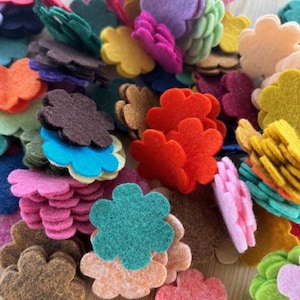 May include: A pile of colorful felt flower shapes in various colors, including red, pink, purple, green, yellow, and brown. The felt flowers are stacked and scattered, creating a vibrant and textured display. These are ideal for crafting and DIY projects.