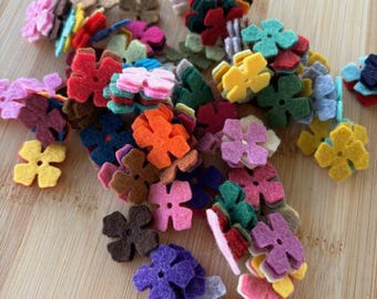 May include: A collection of felt flower shapes in various colors, including red, pink, purple, blue, green, yellow, and brown. The flowers are arranged in a pile on a wooden surface.