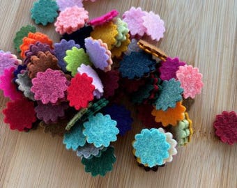 May include: A collection of felt flower shapes in various colors, including red, pink, blue, green, yellow, and brown. The felt flowers are arranged in a pile on a wooden surface.