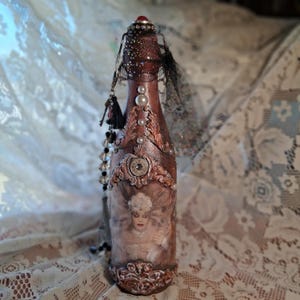 May include: An ornate, decorative bottle with a brown and copper finish. The bottle is adorned with pearls, a red gem, and a black tassel. A vintage image of a woman is featured on the front, adding to the bottle's antique aesthetic.