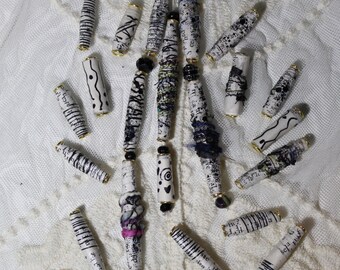 May include: A collection of cylindrical paper beads featuring various black and white patterns, including stripes, dots, and text. Some beads are accented with small dark beads at the ends, while others incorporate subtle hints of purple and pink.