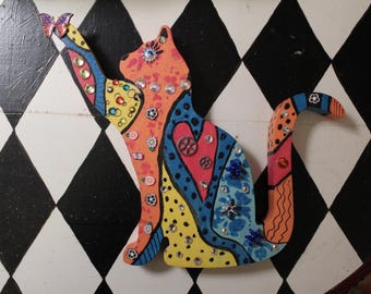May include: A colorful, decorative cat figurine with a raised paw. The wooden cat is painted in orange, yellow, and blue with floral and gem embellishments. A butterfly adorns the raised paw. The cat is set against a black and white diamond pattern.