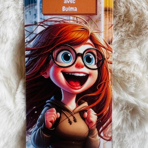 Peut inclure: Un marque-page rectangulaire avec une illustration de dessin animé d'une fille aux cheveux roux, portant des lunettes et souriant largement. Le marque-page porte le texte "Une année avec Bulma" sur une bannière marron. L'arrière-plan montre des horloges et un paysage urbain flou.