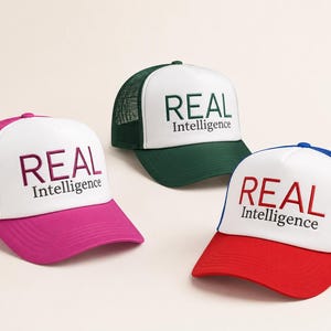 May include: Three trucker caps with white front panels and mesh backs. Each cap features the text REAL Intelligence embroidered on the front. One cap is magenta, one is dark green, and one has a red brim with a blue mesh back and blue accents.