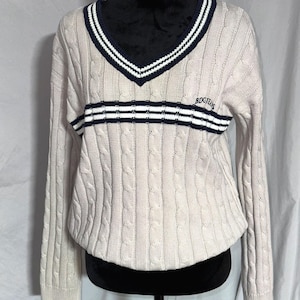 May include: A cream colored cable knit sweater with a navy and white striped v-neck and a navy and white striped band across the chest. The sweater has the text "BDGJEANS" embroidered on the chest.