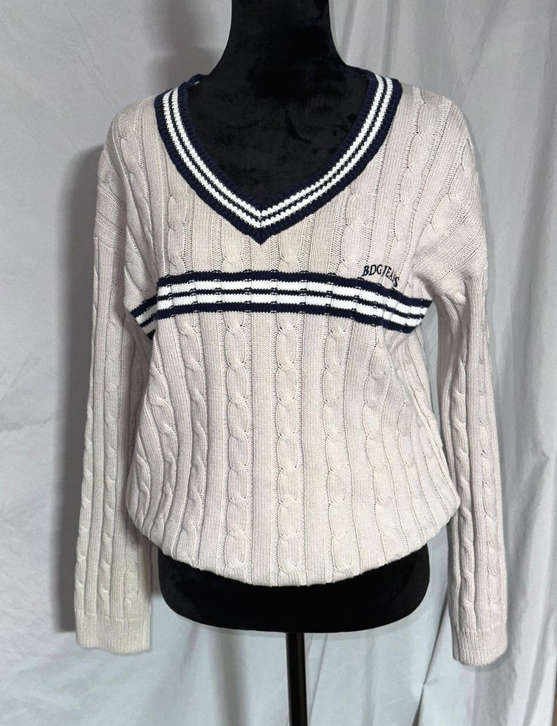 May include: A cream colored cable knit sweater with a navy and white striped v-neck and a navy and white striped band across the chest. The sweater has the text "BDGJEANS" embroidered on the chest.