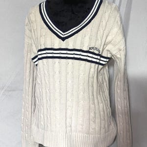 May include: A cream colored cable knit sweater with a navy blue and white striped v-neck collar and a navy blue and white striped band across the chest. The sweater has the text "BDGJEANS" embroidered on the chest.