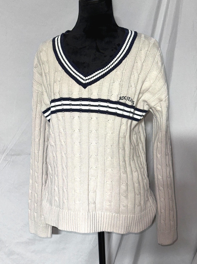 May include: A cream colored cable knit sweater with a navy blue and white striped v-neck collar and a navy blue and white striped band across the chest. The sweater has the text "BDGJEANS" embroidered on the chest.