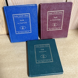 May include: Three vintage pocket-sized opera librettos by Verdi. The books are in blue, burgundy, and teal green. Each book has a gold-colored frame with the title and author's name. The text on the books reads "Pocket Libretto Library" and the titles of the operas.