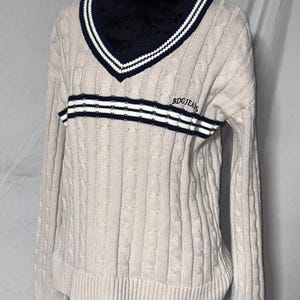 May include: A cream colored cable knit sweater with a navy blue and white striped v-neck collar and a navy blue and white striped band across the chest. The sweater has the text "BDG JEANS" embroidered on the chest.