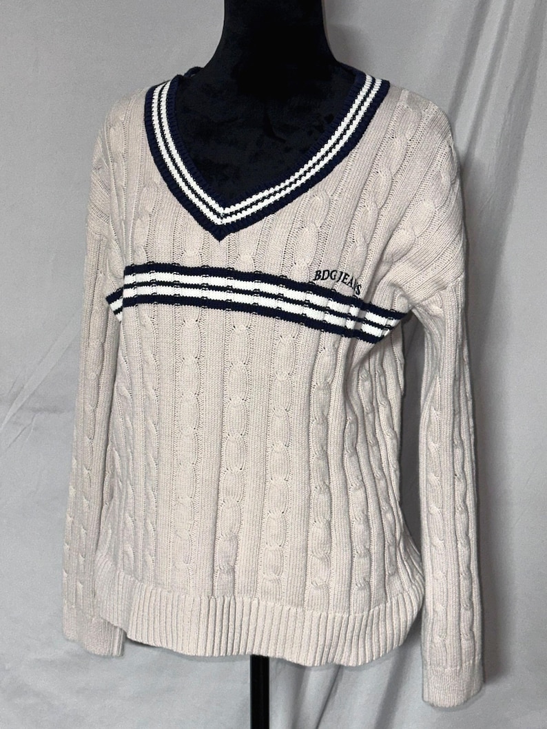 May include: A cream colored cable knit sweater with a navy blue and white striped v-neck collar and a navy blue and white striped band across the chest. The sweater has the text "BDG JEANS" embroidered on the chest.