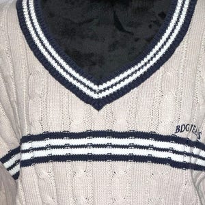 May include: A beige cable knit sweater with a navy blue and white striped v-neck collar and a navy blue and white striped band across the chest. The sweater has the text "BDGJEANS" on the right side of the chest.