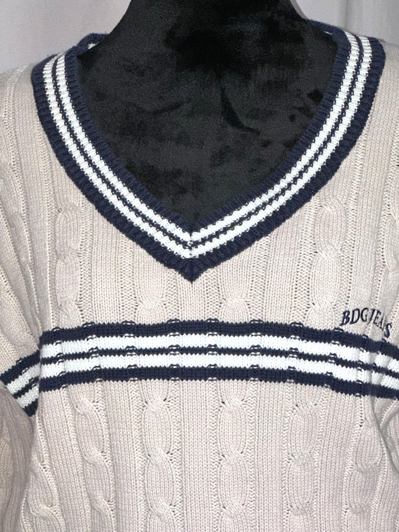 May include: A beige cable knit sweater with a navy blue and white striped v-neck collar and a navy blue and white striped band across the chest. The sweater has the text "BDGJEANS" on the right side of the chest.