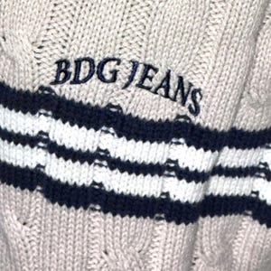 May include: A beige cable knit sweater with a navy blue and white striped band at the bottom. The sweater has the text "BDG JEANS" embroidered in black on the front.