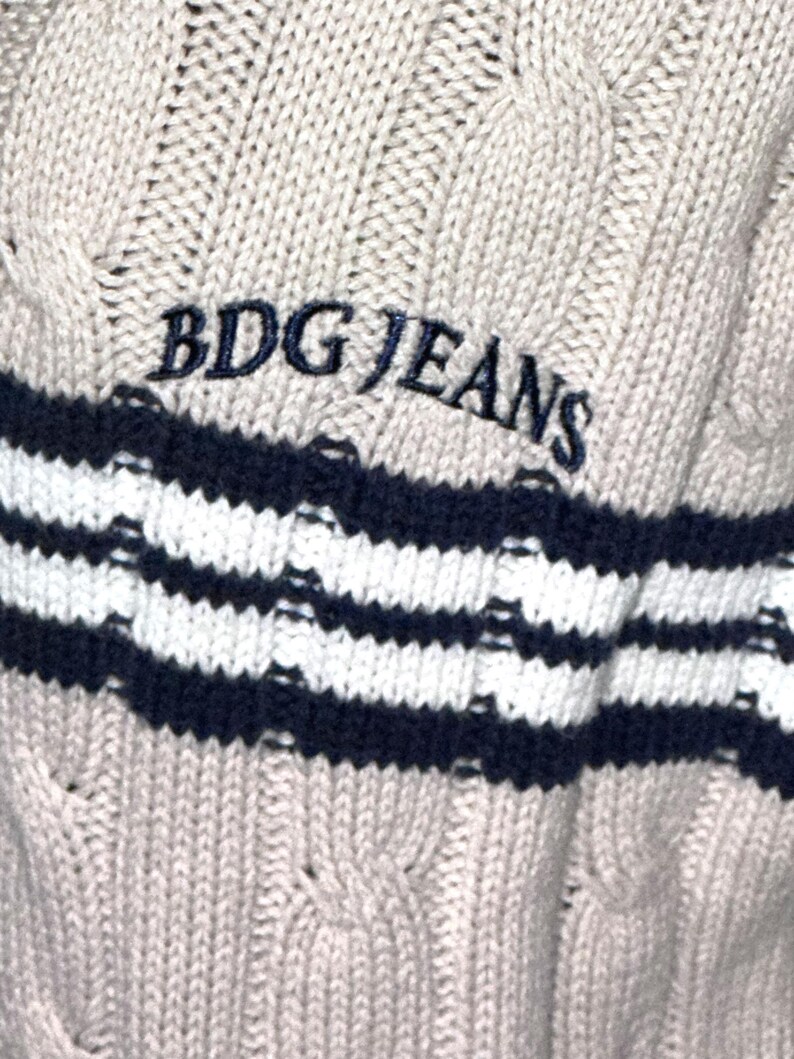 May include: A beige cable knit sweater with a navy blue and white striped band at the bottom. The sweater has the text "BDG JEANS" embroidered in black on the front.
