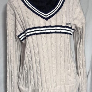 May include: A beige cable knit sweater with a navy blue and white striped collar and a navy blue and white striped band across the chest. The sweater has the word "Bogner" embroidered on the chest.
