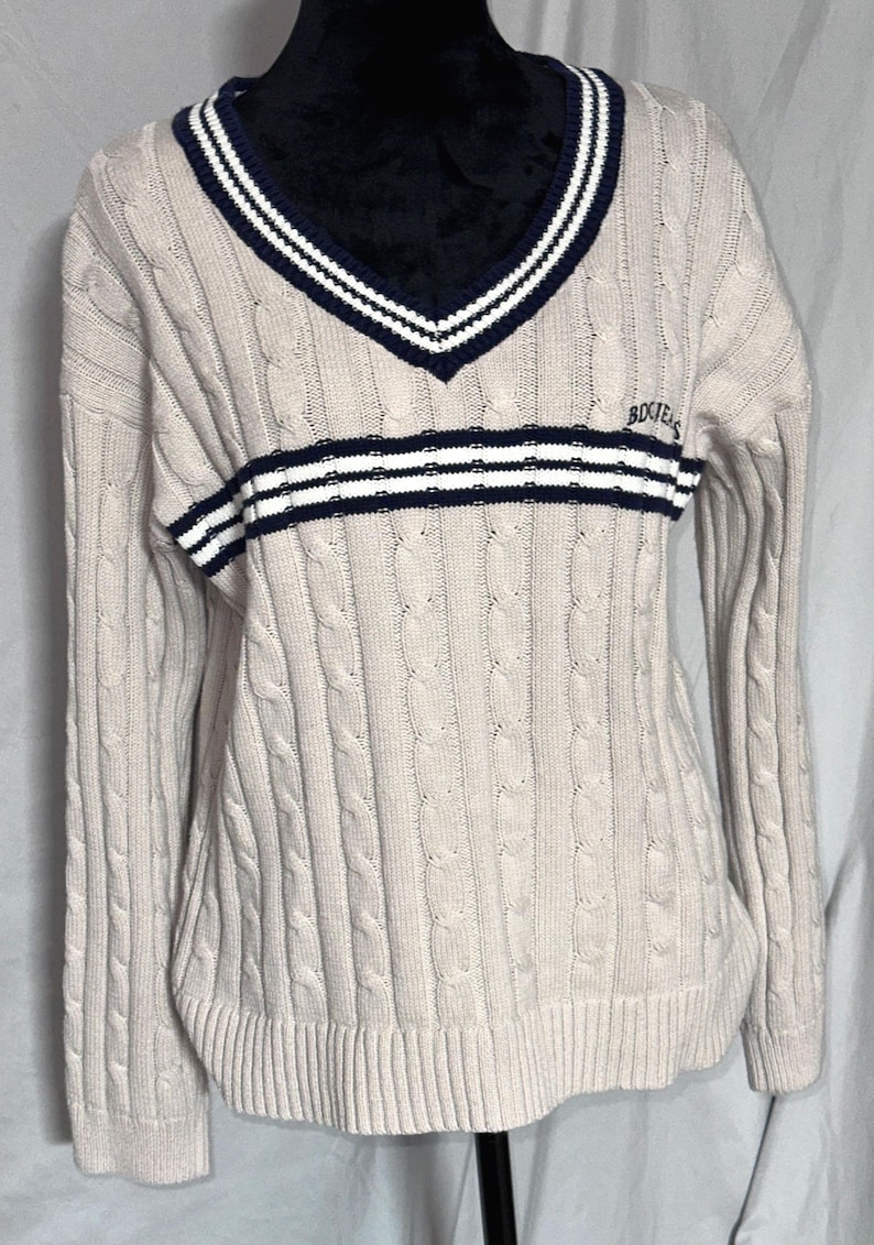 May include: A beige cable knit sweater with a navy blue and white striped collar and a navy blue and white striped band across the chest. The sweater has the word "Bogner" embroidered on the chest.