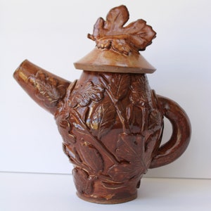 Può includere: Una teiera in ceramica marrone con un design a foglie. La teiera ha un coperchio con una foglia in cima. La teiera è fatta a mano e ha un aspetto rustico.