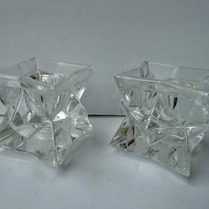 Op de afbeelding: Twee heldere glazen kandelaars met een geometrisch, stervormig ontwerp. Elke houder heeft een vierkante bovenkant en een facetgeslepen, veelzijdige basis. Het transparante glas laat licht door, waardoor een sprankelend effect ontstaat. Deze decoratieve stukken zijn ideaal.