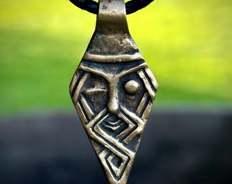 May include: A bronze pendant necklace featuring a stylized face with a beard and a single eye. The pendant is in the shape of a teardrop and has a geometric design.