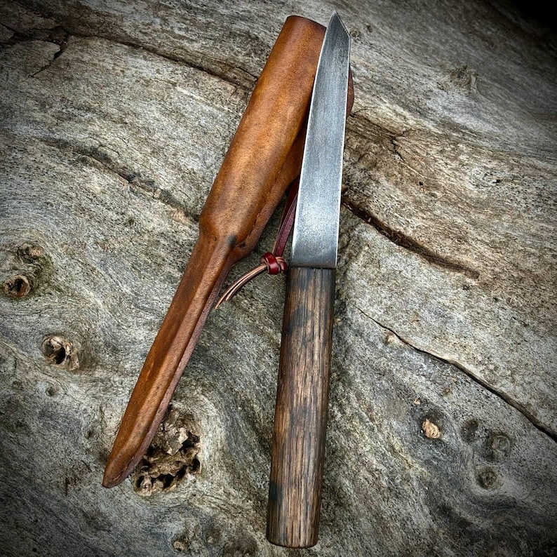 Puede incluir: Un peque&ntilde;o cuchillo forjado a mano con mango de madera y funda de cuero. La hoja es de acero y tiene una ligera curva. La funda es de cuero marr&oacute;n con un detalle de costura roja.