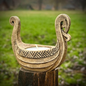 Puede incluir: Un portavelas decorativo con forma de barco vikingo, de color marrón claro. Presenta tallas detalladas y contiene una vela blanca. Se exhibe sobre un poste de madera, sobre un fondo verde borroso. Un objeto decorativo para un ambiente nórdico.