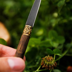 Puede incluir: Un cuchillo esbelto con una hoja de acabado mate oscuro, un mango cil&iacute;ndrico de madera con veta visible y una virola decorativa de lat&oacute;n grabada con un patr&oacute;n floral o de rayos de sol en la uni&oacute;n de la hoja y el mango.