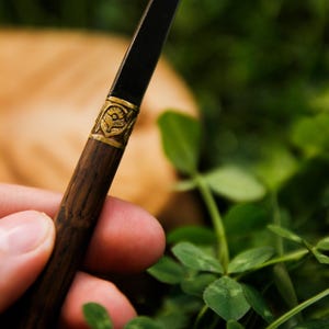 Puede incluir: Un instrumento de escritura con un cuerpo inferior de madera oscura y una secci&oacute;n superior negra, separados por una banda de metal dorado grabada con un motivo floral. El dise&ntilde;o combina texturas de veta de madera natural con detalles met&aacute;licos.