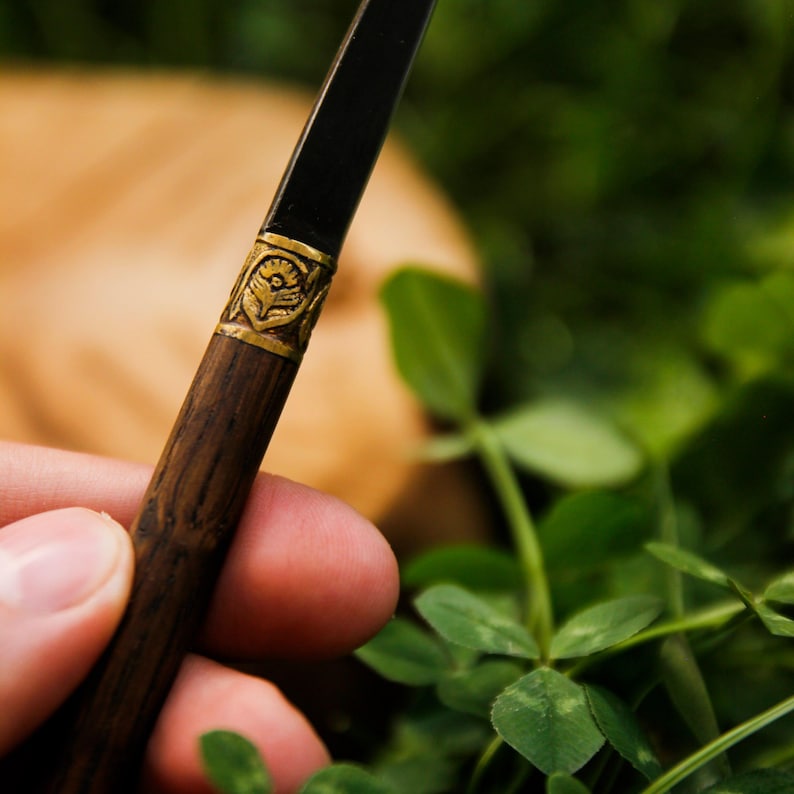 Puede incluir: Un instrumento de escritura con un cuerpo inferior de madera oscura y una secci&oacute;n superior negra, separados por una banda de metal dorado grabada con un motivo floral. El dise&ntilde;o combina texturas de veta de madera natural con detalles met&aacute;licos.