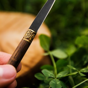Puede incluir: Un abrecartas con una hoja de metal oscura y delgada y un mango cil&iacute;ndrico de madera. El mango presenta una virola decorativa de lat&oacute;n con un patr&oacute;n floral en relieve en la uni&oacute;n donde la hoja se encuentra con la madera, creando contraste.