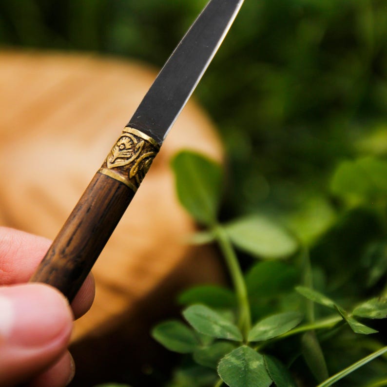 Puede incluir: Un abrecartas con una hoja de metal oscura y delgada y un mango cil&iacute;ndrico de madera. El mango presenta una virola decorativa de lat&oacute;n con un patr&oacute;n floral en relieve en la uni&oacute;n donde la hoja se encuentra con la madera, creando contraste.