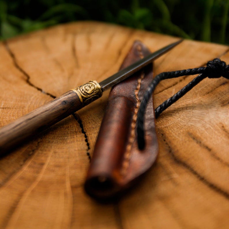 Puede incluir: Una hoja de metal delgada con un mango de madera oscura que presenta un refuerzo de lat&oacute;n decorativo con un dise&ntilde;o circular en relieve. Junto a la hoja hay una funda de cuero marr&oacute;n con costuras visibles y un cord&oacute;n negro adjunto.