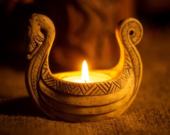 May include: A beige ceramic candle holder shaped like a Viking longship with a carved dragon head at the prow and a curled tail at the stern. The hull features etched geometric patterns and a lit tea light candle sits in the center.
