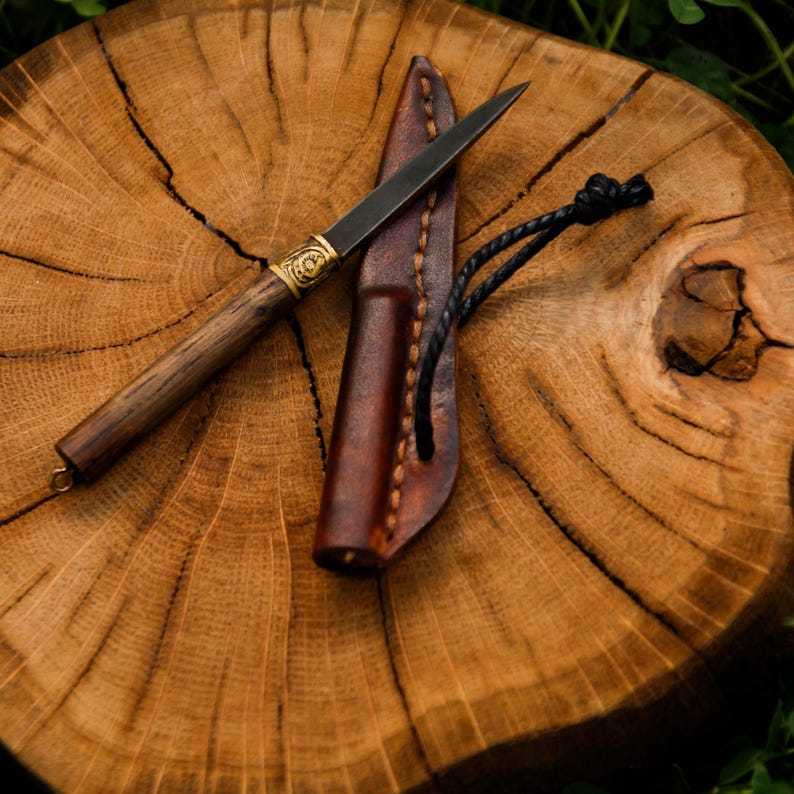 Puede incluir: Un cuchillo peque&ntilde;o con hoja de metal oscuro, mango de madera y virola de lat&oacute;n con un dise&ntilde;o grabado. El cuchillo viene acompa&ntilde;ado de una funda de cuero marr&oacute;n con costuras visibles y un cord&oacute;n negro unido al lateral para poder colgarlo.
