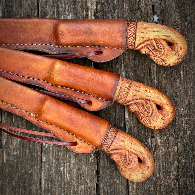 Op de afbeelding: Drie handgemaakte messen met leren scheden. De scheden zijn warmbruin en de handvatten zijn van gesneden hout met ingewikkelde patronen. De messen liggen op een verweerd houten oppervlak.